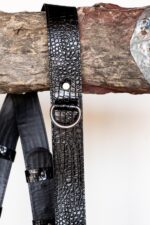 Extreme close-up of a single shoulder strap draped over a rustic wooden branch, highlighting the detailed glossy black crocodile-embossed leather texture, a silver rivet, and the D-ring attachment loop.