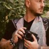 Photographer wearing the Black Croc leather camera harness over a plaid blazer and black t-shirt, holding a Fujifilm camera, with the crocodile-embossed straps and silver D-ring hardware visible on the shoulders.