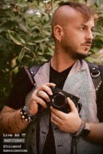 Photographer wearing the Black Croc leather camera harness over a plaid blazer and black t-shirt, holding a Fujifilm camera, with the crocodile-embossed straps and silver D-ring hardware visible on the shoulders.