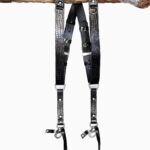 Full-length view of the Black Croc leather dual camera harness hanging from a rustic wooden branch, showing the X-shaped crossback design with black leather backing, black-tone buckles and keeper loops, silver D-ring connectors, and snap-hook attachments at the bottom.