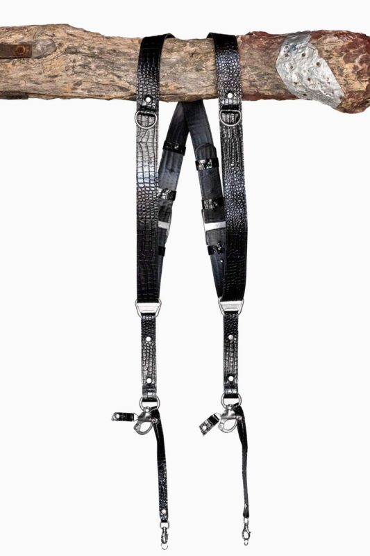 Full-length view of the Black Croc leather dual camera harness hanging from a rustic wooden branch, showing the X-shaped crossback design with black leather backing, black-tone buckles and keeper loops, silver D-ring connectors, and snap-hook attachments at the bottom.