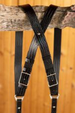 Close-up of the X-shaped crossback junction of the Black Croc harness hanging from a wooden branch against a warm wood-paneled background, highlighting the glossy crocodile texture, center rivet, dual silver buckles, and black leather backing straps.
