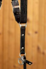 Detail view of a single strap end showing the "GENUINESTRAP" branded triangular D-ring connector, glossy black crocodile-embossed leather with silver rivets, O-ring, and marine-grade snap hook with a croc leather safety tab, against a warm wood background.