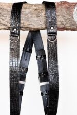Close-up of both shoulder straps hanging from a rustic wooden branch, showcasing the glossy black crocodile-embossed leather texture, silver rivet-mounted D-rings, black leather backing with keeper loops, and the branded silver slide buckles.