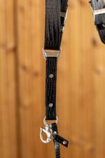 Detail view of a single strap end showing the branded triangular D-ring connector, crocodile-embossed black leather with silver rivets, O-ring, and snap-hook camera attachment with a croc leather tab, against a warm wood-paneled background.