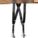 Black leather dual camera harness hanging from a rustic wooden beam with adjustable straps and silver hardware.