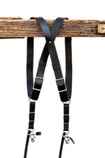 Black leather dual camera harness hanging from a rustic wooden beam with adjustable straps and silver hardware.