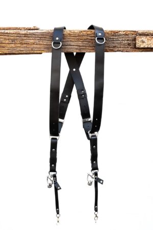 Black leather dual camera harness hanging from a rustic wooden beam with adjustable straps and silver hardware.