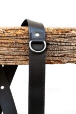 Close-up of a black leather shoulder strap with silver D-ring hanging over a weathered wooden beam.