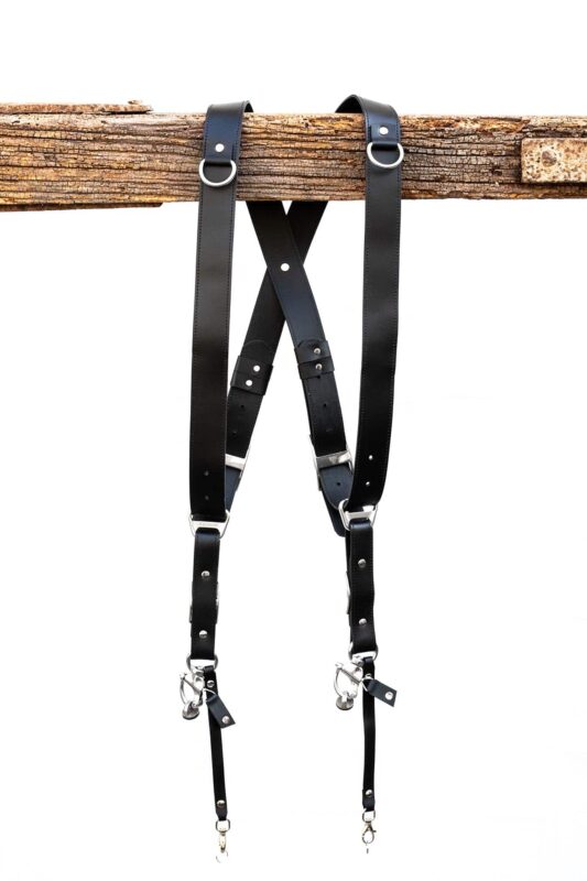 Black leather dual camera harness hanging from a rustic wooden beam with adjustable straps and silver hardware.