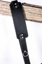 Black leather camera strap close-up with padded shoulder section, stitched edges, silver rivets, and metal ring detail hanging over a rustic wooden beam on a light gray background.
