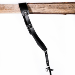 Full-length black leather camera strap with padded shoulder pad, adjustable strap holes, silver metal hardware, and clasp attachment hanging from a rustic wooden beam on a light gray background.