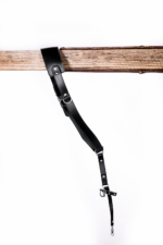 Full-length black leather camera strap with padded shoulder pad, adjustable strap holes, silver metal hardware, and clasp attachment hanging from a rustic wooden beam on a light gray background.