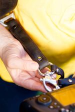 Close-up of a hand attaching a camera to a dark leather strap with a metal swivel clasp.