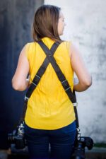 Back view of a woman wearing a dark leather dual camera harness with cameras attached on both sides.