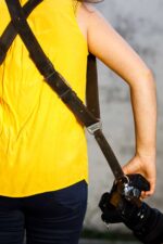 Woman wearing a dark leather dual camera strap over a bright yellow sleeveless top, holding a camera at her side.