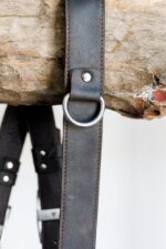 Close-up of a dark leather camera strap with stitched edges and a metal D-ring hanging over weathered wood.