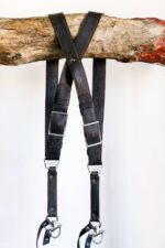 Full view of a dark leather dual camera harness with adjustable straps and metal clasps hanging from a wooden branch.