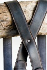 Close-up of crossed dark leather shoulder straps with stitching detail draped over a rustic wooden branch.