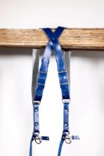 Blue Shanty leather camera harness displayed flat on weathered wood beam, showing full X-harness design with royal blue shoulder straps, grey center strap with navy leather accent patches, metal D-ring attachments, and carabiner clips