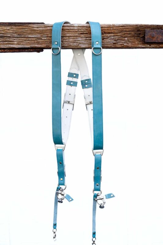 Full display of Blue Sirocco leather camera harness showing complete X-design with turquoise shoulder straps, white center X-strap with teal leather accent patches, metal square connectors, adjustable leather segments, and dual metal carabiner clips with leather tags