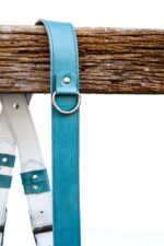 Close-up detail of Blue Sirocco harness shoulder strap showing metal D-ring connector, turquoise leather with white stitch detailing, and center white strap with teal leather accent patch visible
