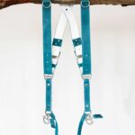Full front product display of Blue Vintage leather camera harness on weathered wood showing dual teal leather shoulder straps with metal D-rings, white center X-strap with teal and navy accent bands, multiple metal square connectors, white stitching, and carabiner clips with leather attachment tags