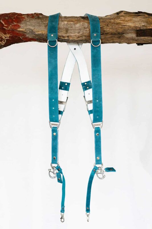 Full front product display of Blue Vintage leather camera harness on weathered wood showing dual teal leather shoulder straps with metal D-rings, white center X-strap with teal and navy accent bands, multiple metal square connectors, white stitching, and carabiner clips with leather attachment tags