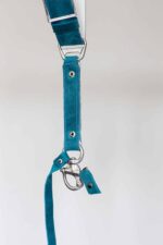 Vertical detail of Blue Vintage harness lower strap showing teal leather with white stitching, metal connector hardware, white metal trim, rivets, and carabiner clip attachment with leather tag
