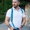 Man wearing Blue Vintage leather camera harness in white short-sleeve polo shirt and black pants, holding DSLR camera, standing against ivy-covered stone wall background with side profile view