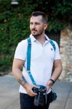 Man wearing Blue Vintage leather camera harness in white short-sleeve polo shirt and black pants, holding DSLR camera, standing against ivy-covered stone wall background with side profile view