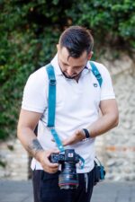 Man in white polo shirt looking down at DSLR camera suspended on Blue Vintage teal leather harness with metal D-ring connector and white metal hardware, stone wall setting
