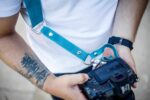 Close-up detail of Blue Vintage harness showing teal leather shoulder strap with white metal D-ring, white center strap connector band with white rivets, worn against white shirt while holding black DSLR