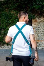 Back view of man wearing complete Blue Vintage camera harness with dual teal leather shoulder straps forming X-design, white center strap with teal accent bands, metal square connectors, supporting two DSLR cameras, ivy-covered wall background