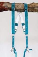 Blue Vintage leather camera harness displayed on weathered wood beam showing full X-harness design with dual teal shoulder straps with metal D-rings, white center cross-strap with teal and navy accent bands, white stitching throughout
