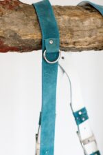 Close-up detail of Blue Vintage harness shoulder strap showing teal vintage leather with white stitching, metal D-ring connector, white center strap visible with layered accent band detail