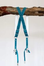 Complete back display of Blue Vintage harness on aged wood showing dual teal shoulder straps with metal D-rings, white center X-strap with teal and darker navy accent bands, metal square connectors, white stitching, and carabiner clips with leather tags