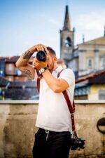 Man using a camera while wearing a dark brown leather dual camera harness, outdoor lifestyle shot.