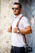 Man wearing a dark brown leather dual camera harness, adjusting the straps on his chest.