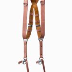 Brown Siena leather camera harness displayed on light wood beam, showing full X-harness design with mauve-pink leather shoulder straps, rich brown center strap with tan accent bands, metal D-ring connectors, and carabiner clips