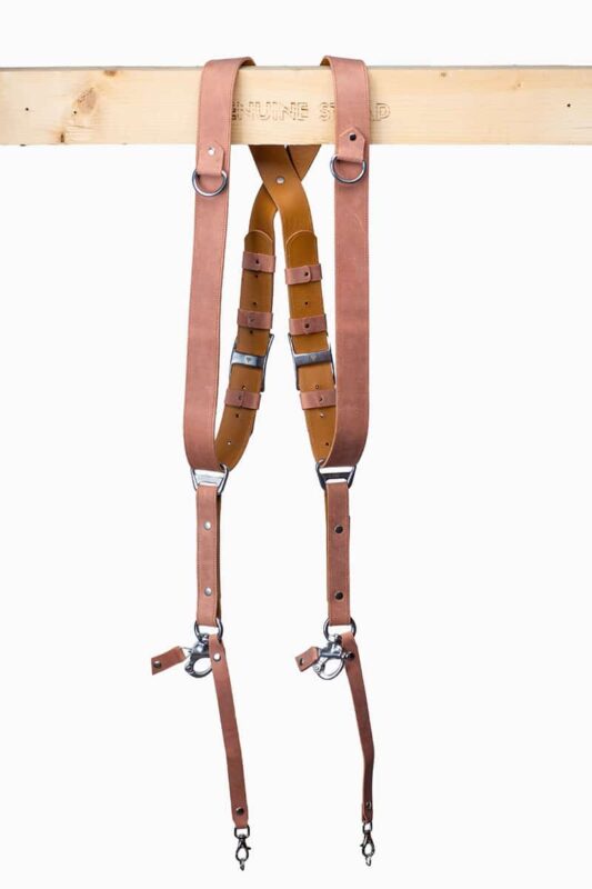 Brown Siena leather camera harness displayed on light wood beam, showing full X-harness design with mauve-pink leather shoulder straps, rich brown center strap with tan accent bands, metal D-ring connectors, and carabiner clips