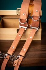 Brown Siena leather camera harness draped over branded wooden box against teal background, displaying peachy-tan shoulder straps, brown center strap with metal hardware, and complete X-harness structure