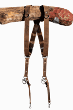 Full-length front view of the Brown Vintage leather dual camera harness hanging from a rustic wooden branch, showing the complete harness with distressed tan leather straps, dark brown crossback, D-ring shoulder attachments, antique-finish buckles, branded triangular connectors, and snap-hook camera attachments with leather safety tabs at the bottom.