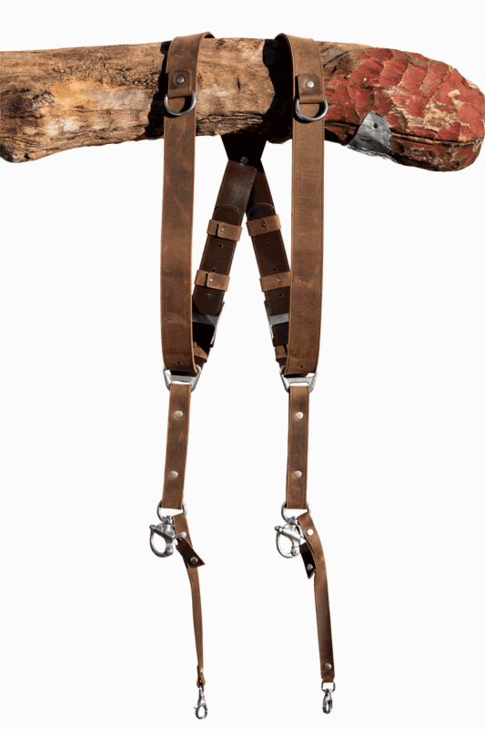 Full-length front view of the Brown Vintage leather dual camera harness hanging from a rustic wooden branch, showing the complete harness with distressed tan leather straps, dark brown crossback, D-ring shoulder attachments, antique-finish buckles, branded triangular connectors, and snap-hook camera attachments with leather safety tabs at the bottom.