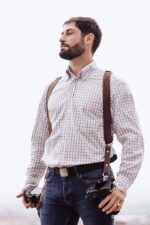 Photographer wearing the Brown Vintage leather camera harness over a checked button-down shirt, shown from the front with two cameras hanging at hip level, demonstrating how the harness sits comfortably on the shoulders during a shoot.