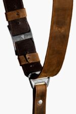 Close-up detail of the Brown Vintage leather camera harness showing the eagle-embossed antique-finish slide buckle, dark brown leather keeper loops, distressed tan leather strap, adjustment holes, and the "GENUINESTRAP" branded triangular D-ring connector.