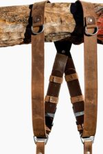 Close-up of both shoulder straps hanging from a rustic wooden branch, showcasing the distressed tan leather with D-ring attachment loops, dark brown leather crossback with stitched rounded tabs, brown keeper loops, and antique-finish buckles with branded triangular connectors below.