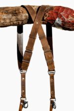 Close-up of the X-shaped crossback junction of the Brown Vintage harness hanging from a rustic wooden branch, showing the distressed tan leather crossing with a center rivet, brown leather keeper loops, dual antique-finish square buckles, and branded triangular D-ring connectors.