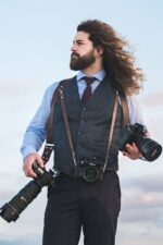 Photographer wearing the Brown Vintage leather camera harness over a tweed waistcoat, blue shirt, and polka-dot tie, carrying two Nikon cameras at hip level with hair blowing in the wind, demonstrating the harness in use during a shoot.