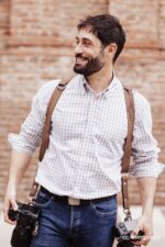 Photographer wearing the Brown Vintage leather camera harness over a checked button-down shirt, smiling while holding two cameras at his sides, with the brown leather straps and D-ring shoulder attachments visible against a brick wall backdrop.